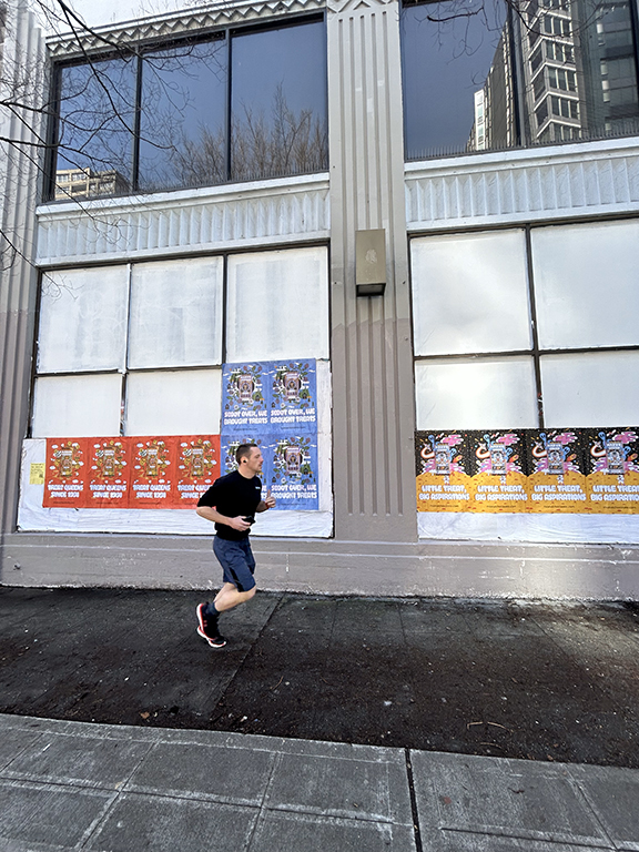 Man jogging past a wall covered with colorful wheatpaste posters advertising various events and campaigns, showcasing urban guerrilla marketing strategies.