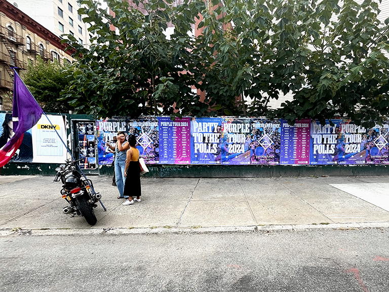Two people standing near a motorcycle in front of a wall covered with vibrant wheatpaste posters for the "Party to the Polls 2024" campaign, showcasing event details and dates in an urban setting.