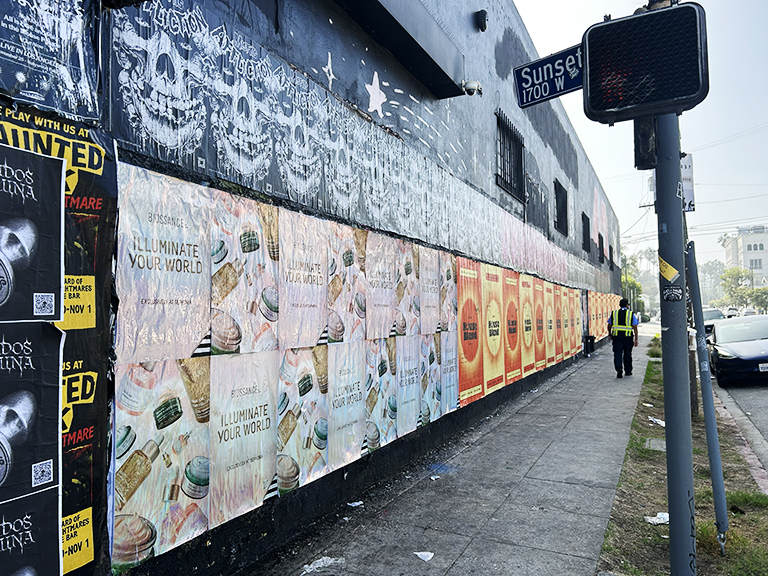 Wheatpaste advertising campaign featuring multiple colorful posters for Biossance's "Illuminate Your World" on a wall in an urban setting, with pedestrians and street signage visible.
