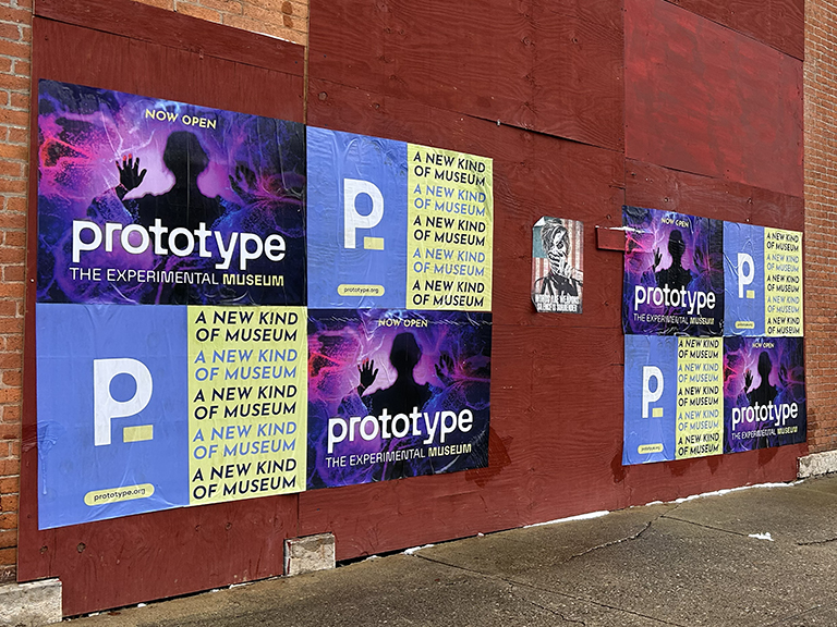 Wheatpaste posters advertising "Prototype - The Experimental Museum," featuring bold graphics and text, displayed on a brick wall in an urban setting, promoting a new kind of museum experience.