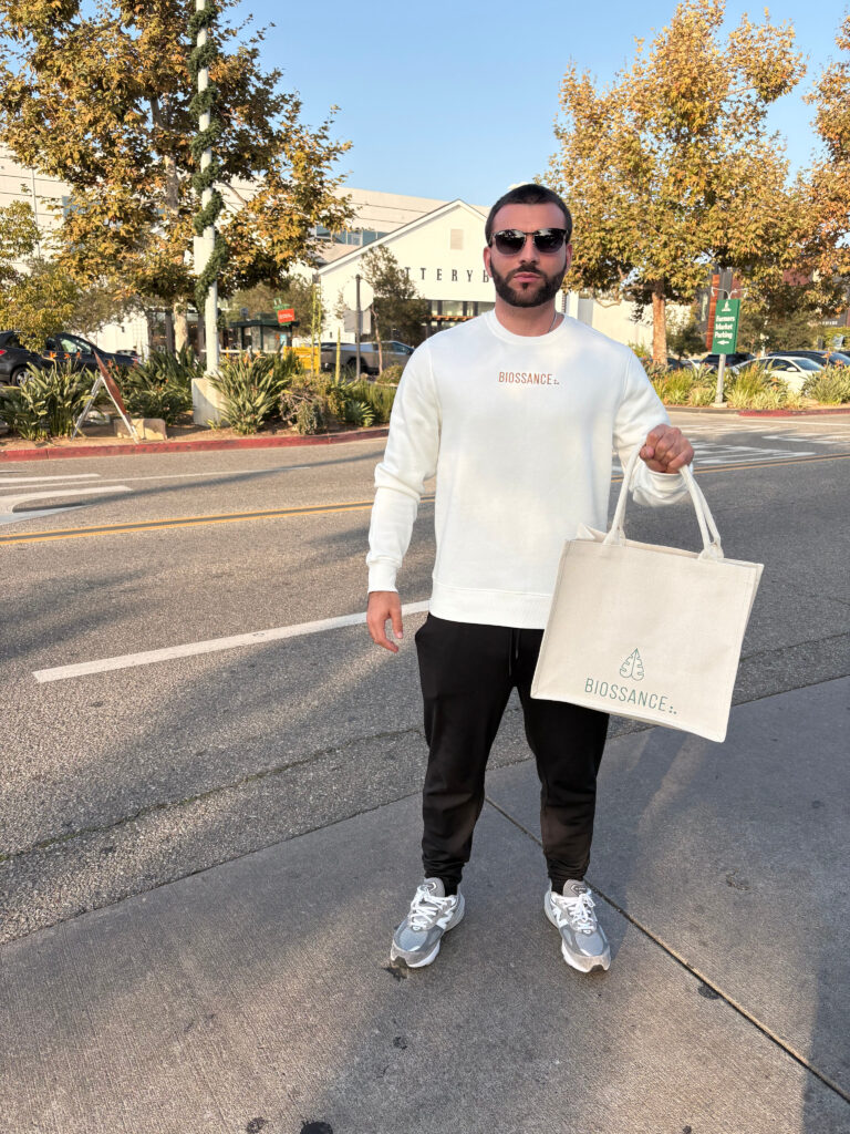 Man holding a Biossance tote bag, wearing branded apparel, standing on a sidewalk with trees and a retail building in the background, promoting Biossance skincare products.