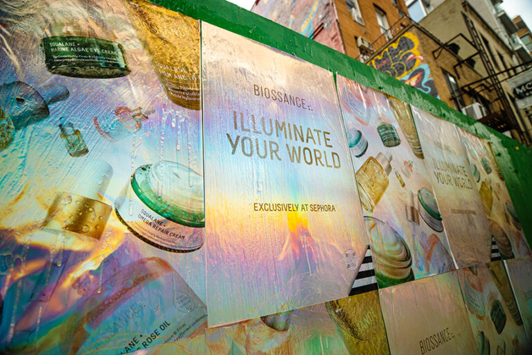 Holographic posters promoting Biossance's "Illuminate Your World" campaign, featuring skincare products and emphasizing exclusivity at Sephora, displayed on an urban wall.