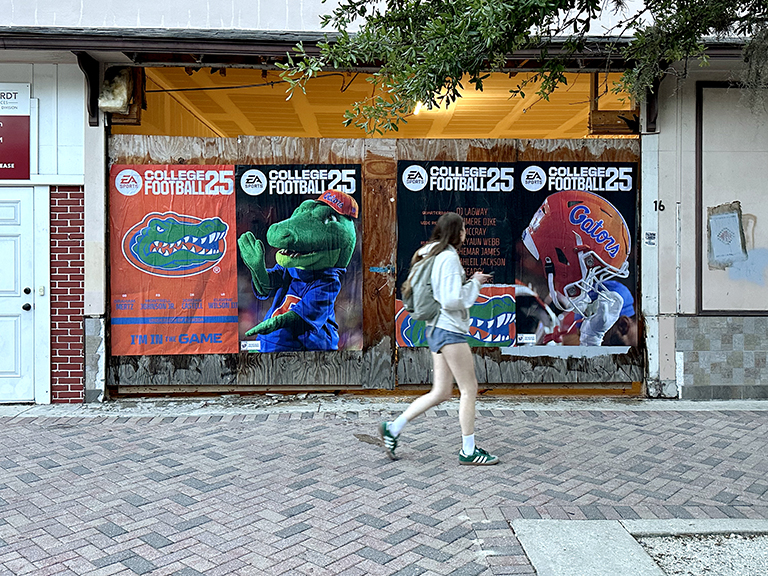 Student walking past promotional banners for College Football 25, featuring Gators mascot and game branding, emphasizing college marketing engagement.