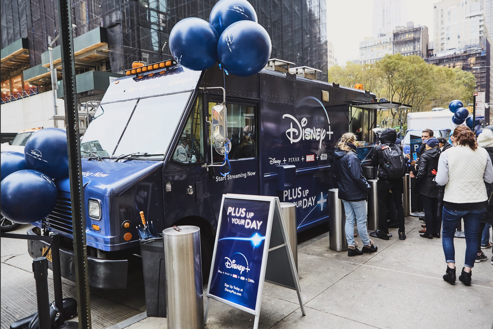 Disney+ branded food truck with promotional balloons and a crowd of people engaging in a pop-up activation event, featuring a sign that reads "PLUS UP YOUR DAY."