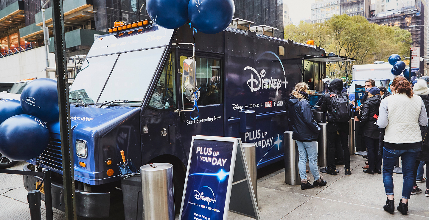 Disney+ promotional food truck attracting a crowd with blue balloons and signage reading "Plus Up Your Day," showcasing guerrilla marketing strategy for high visibility and engagement.