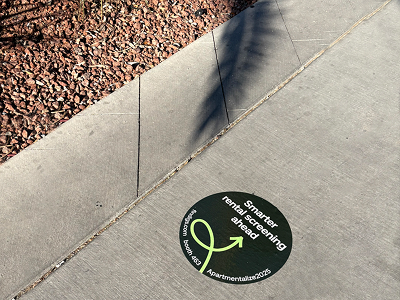 Circular floor graphic with text "Smarter rental screening ahead" and an arrow, promoting apartment rental services, on a concrete sidewalk among pebbles.
