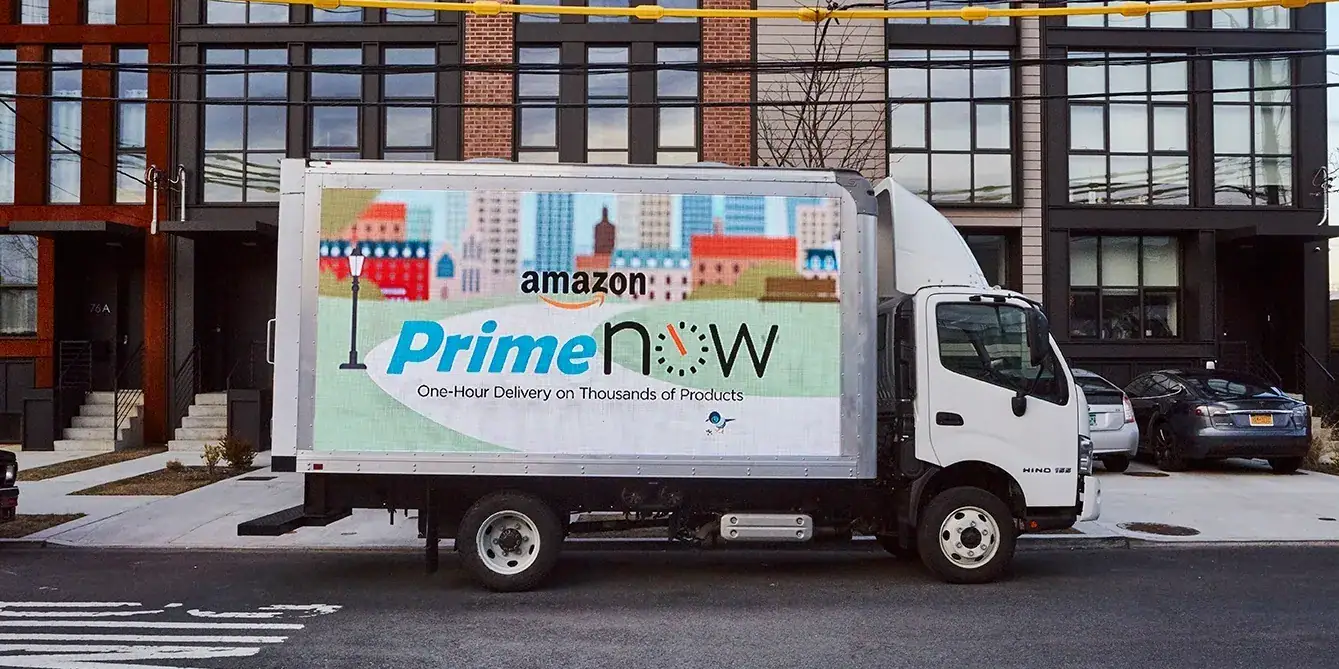 Mobile LED billboard truck showcasing Amazon Prime Now advertisement for one-hour delivery, parked in an urban setting with modern buildings in the background.