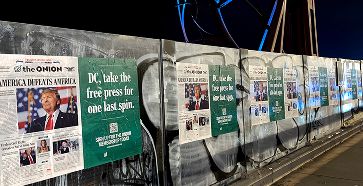 Billboard featuring satirical newspaper headlines from The Onion, including "America Defeats America" and "DC, take the free press for one last spin," displayed on a graffiti-covered wall, emphasizing grassroots political messaging and community engagement.