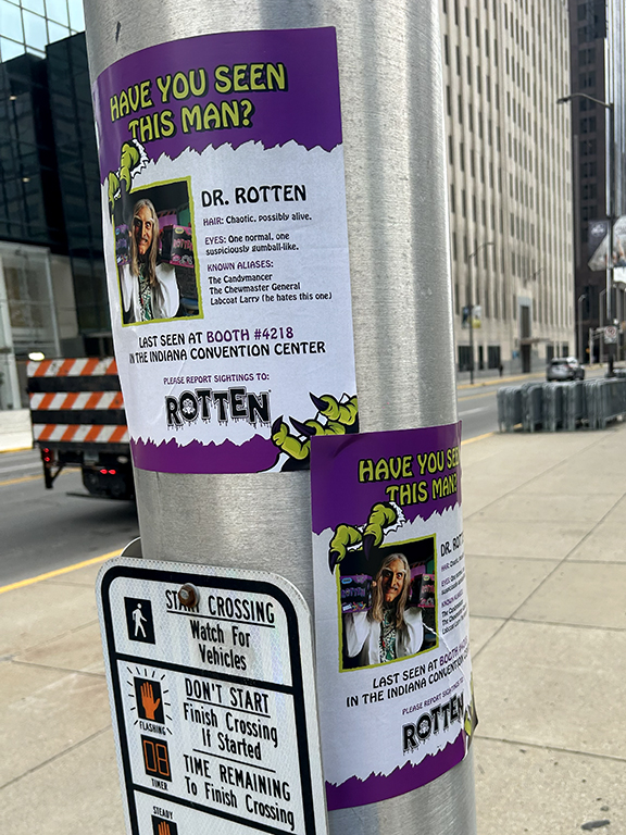 Posters on a street pole advertising a fictional character, Dr. Rotten, with a cartoonish design, featuring a photo, description, and details about his last known location at a convention center, highlighting the playful and engaging nature of convention marketing.