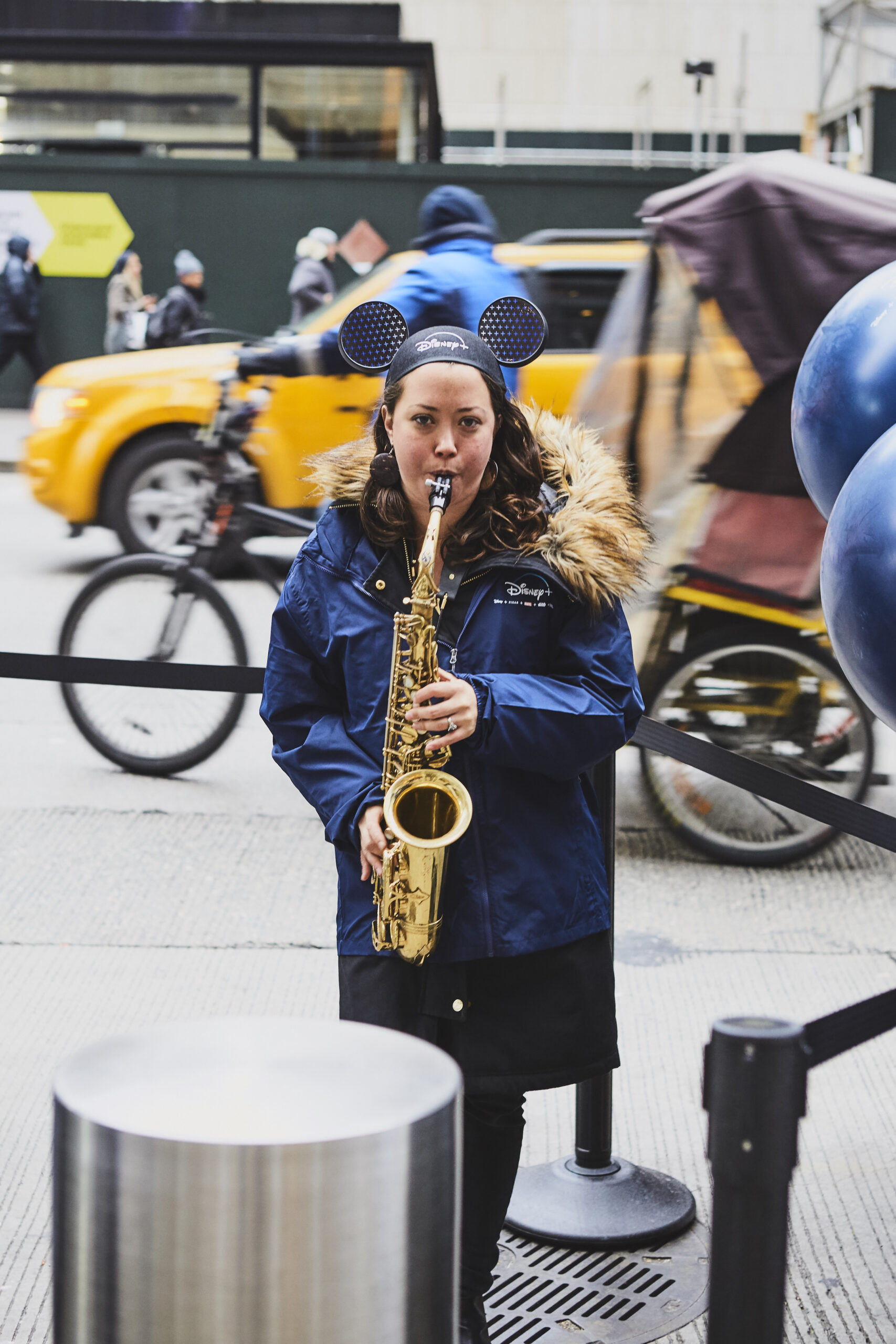 Musician in Disney-themed attire playing saxophone on a busy street, surrounded by pedestrians and yellow taxi cabs, embodying vibrant urban engagement and creative marketing stunts.