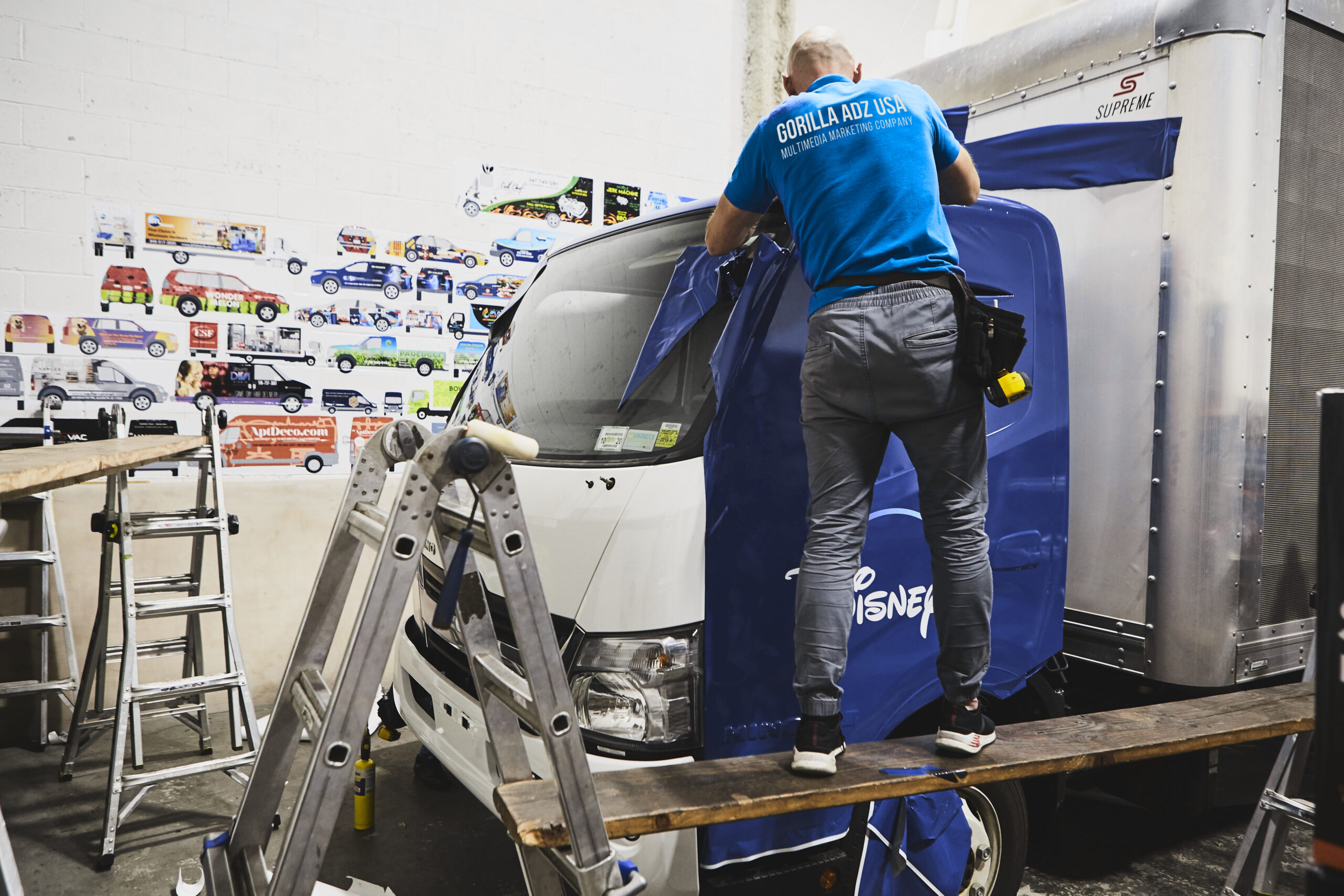 Man applying a blue vinyl wrap to a commercial vehicle in a workshop, showcasing vehicle branding and advertising services.