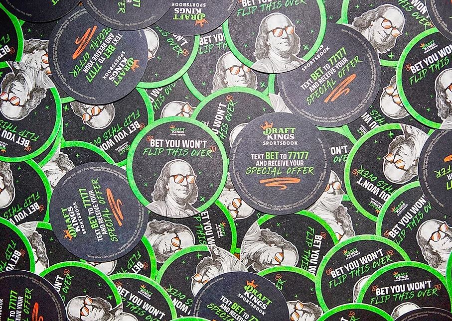 Branded beverage coasters featuring Draft Kings logo and promotional text, emphasizing a special offer and encouraging interaction through a betting message, suitable for advertising in social settings.