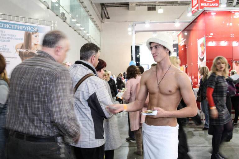 Male distributor in a towel and hat handing out flyers at a busy event, engaging with attendees in a marketing campaign setting.