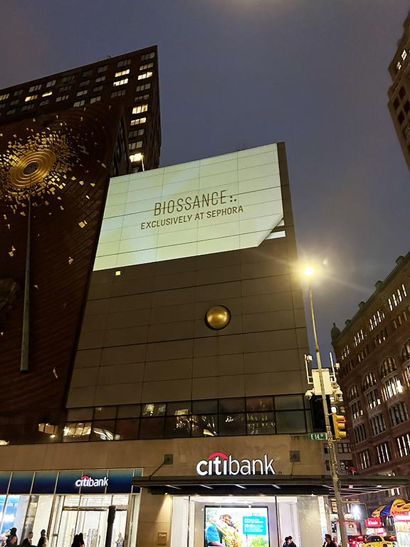 Projection advertisement for Biossance exclusively at Sephora on a building facade near a Citibank storefront, illuminated at night.