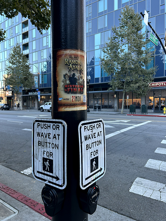 Street pole with a promotional poster for Ramon Ayala's concert, featuring event details, alongside pedestrian crossing buttons for accessibility, set against an urban backdrop with modern buildings.