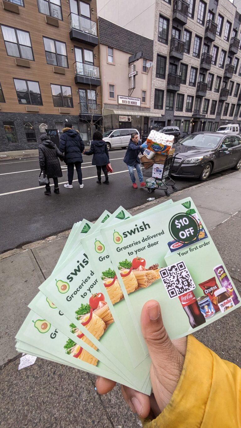 Hand holding flyers for Swish grocery delivery, featuring promotional offer, with people in the background engaging in flyer distribution on a city street.