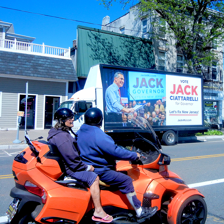 Mobile billboard promoting Jack Ciattarelli for Governor, featuring campaign message and image of Ciattarelli, with two riders on a motorcycle in foreground, set in a busy street context.