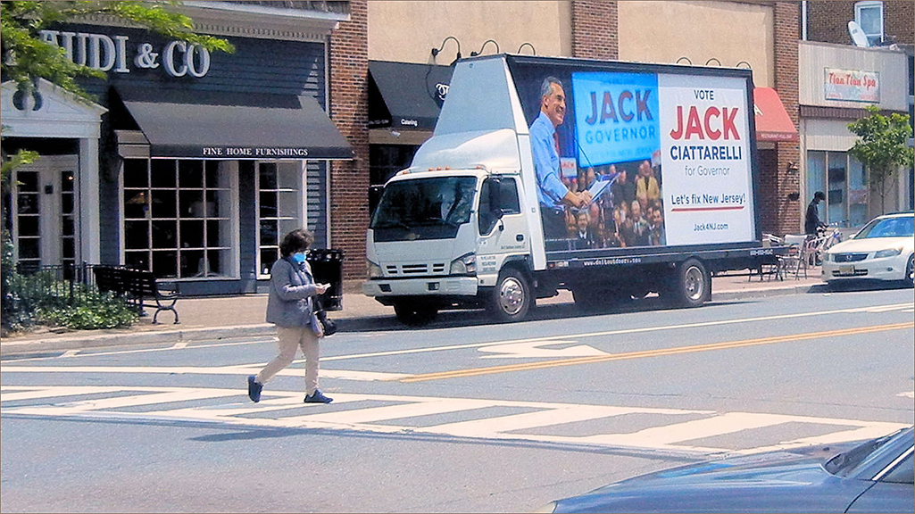 Jack Ciattarelli mobile billboard truck promoting gubernatorial campaign, featuring large graphics and messaging, positioned in a busy town center with pedestrians and retail shops nearby.