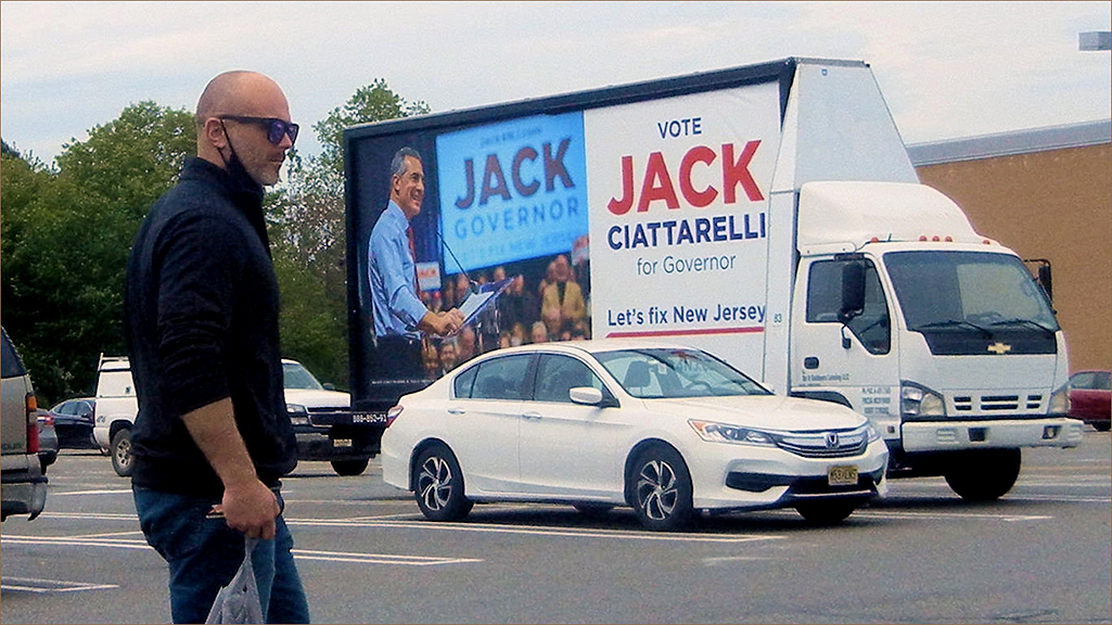 Man walking past a mobile billboard advertising Jack Ciattarelli for Governor, featuring a campaign slogan and image, in a parking lot setting.