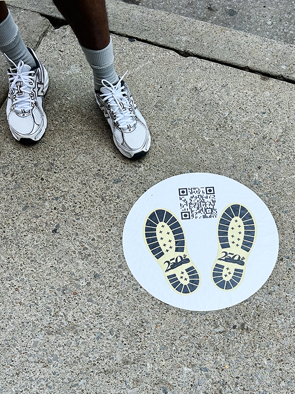 Person standing next to a circular floor graphic featuring shoe prints and a QR code, promoting interactive engagement in a community marketing campaign.