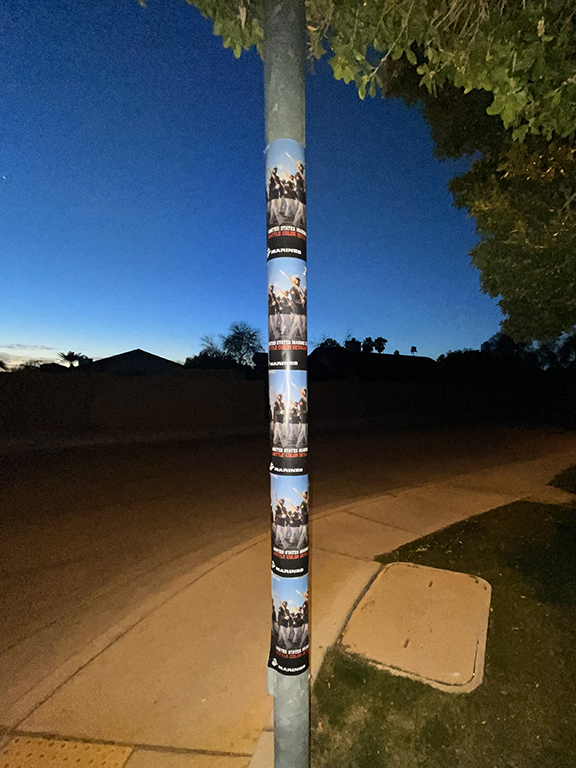 USMC recruitment wild posting on a street pole, featuring multiple posters with bold graphics and messaging, set in a suburban environment during dusk to enhance visibility among high-school students and commuters.