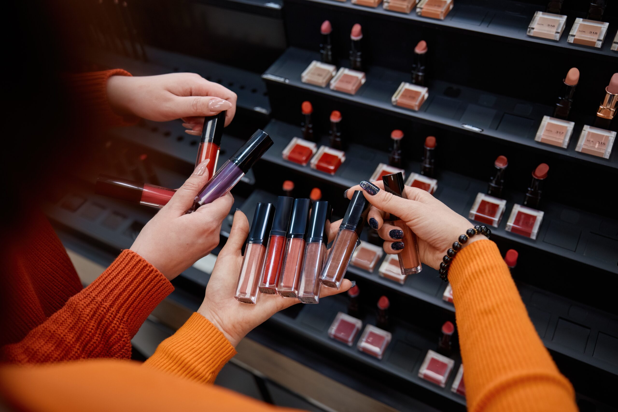 Two individuals selecting various lip glosses from a display, showcasing a range of colors and textures in a retail environment, emphasizing product engagement and sampling.