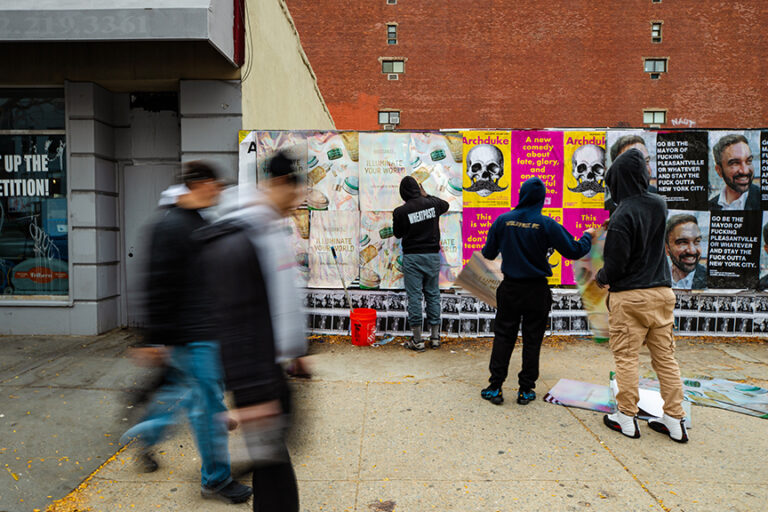 Individuals applying vibrant wheatpaste posters on a city wall, showcasing various designs, including promotional art for campaigns, amidst a bustling urban environment.