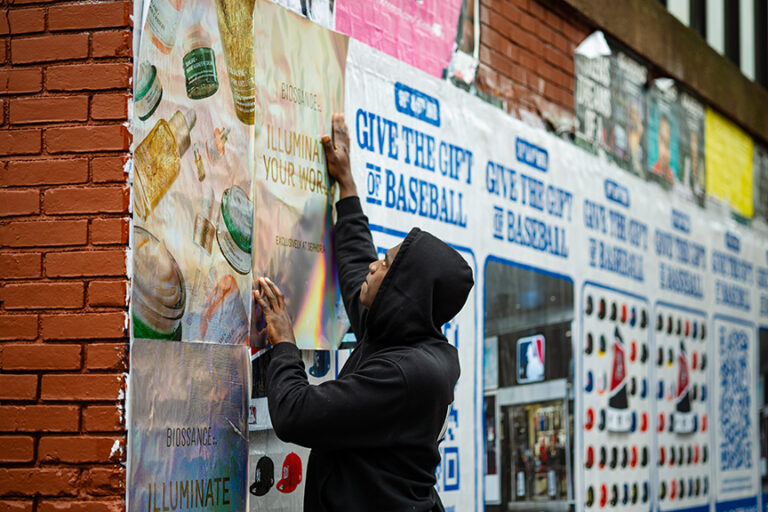 Person applying a Biossance wheatpaste poster promoting the "Illuminate Your World" campaign on a brick wall, surrounded by various other advertisements.