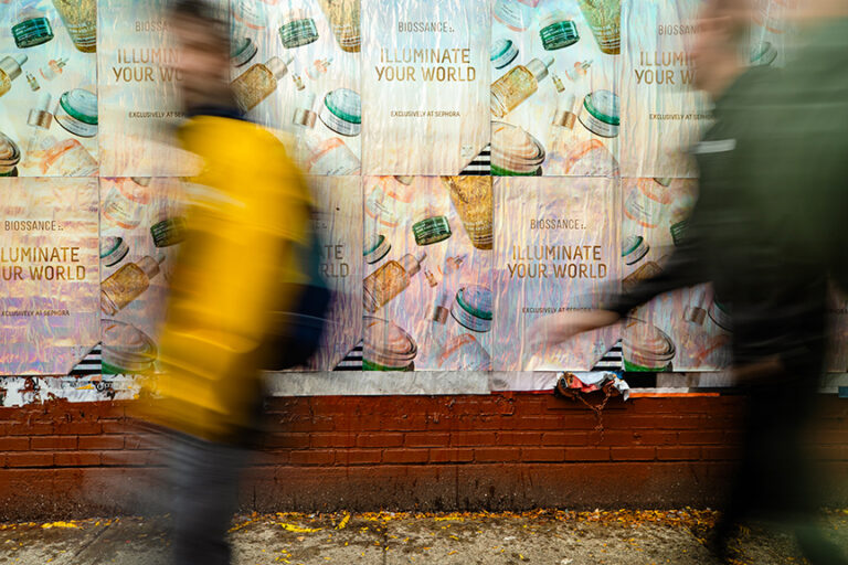 Holographic wheatpaste posters for Biossance "Illuminate Your World" campaign on urban wall, with pedestrians in motion.