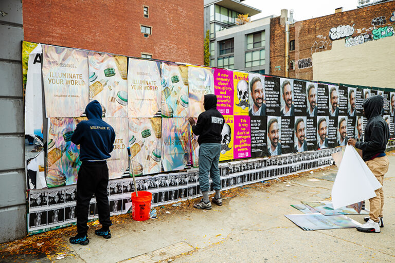 Two individuals applying wheatpaste posters featuring the "Illuminate Your World" campaign on an urban wall, surrounded by various other promotional materials, showcasing guerrilla marketing techniques.
