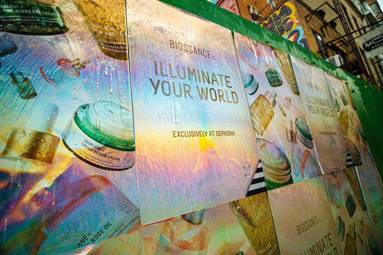 Custom-printed holographic wheatpaste posters for Biossance "Illuminate Your World" campaign displayed on an urban wall, showcasing innovative advertising techniques by American Guerrilla Marketing.