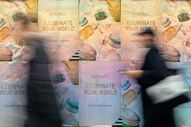 Biossance "Illuminate Your World" wheatpaste posters displayed in urban setting, featuring vibrant skincare product visuals, with blurred figures passing by, highlighting guerrilla marketing effectiveness.