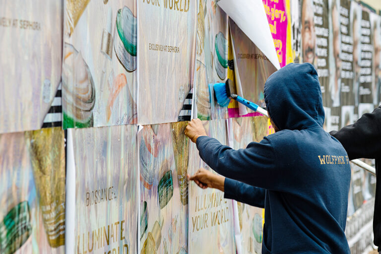 Person applying wheatpaste posters for Biossance "Illuminate Your World" campaign, showcasing guerrilla marketing techniques in urban setting.