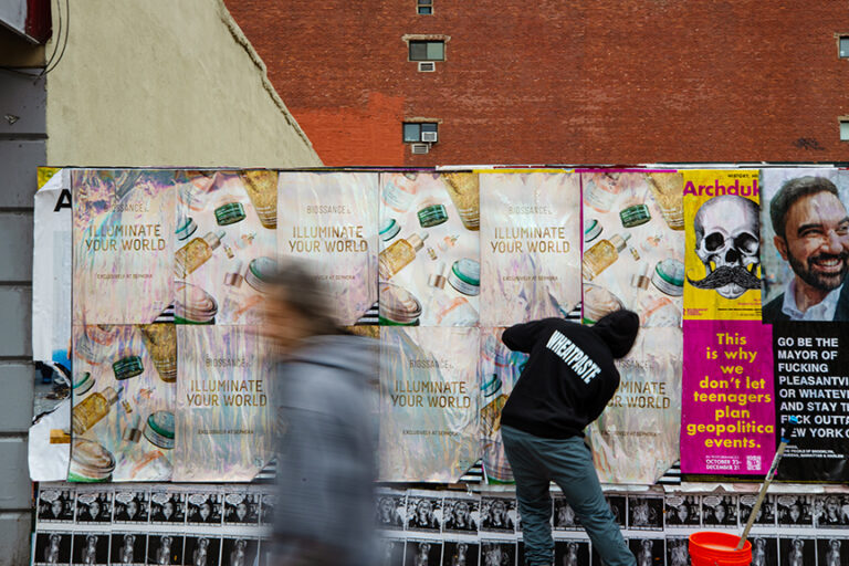 Person pasting wheat posters featuring &ldquo;Illuminate Your World&rdquo; campaign in urban setting, showcasing guerrilla marketing technique by American Guerrilla Marketing.
