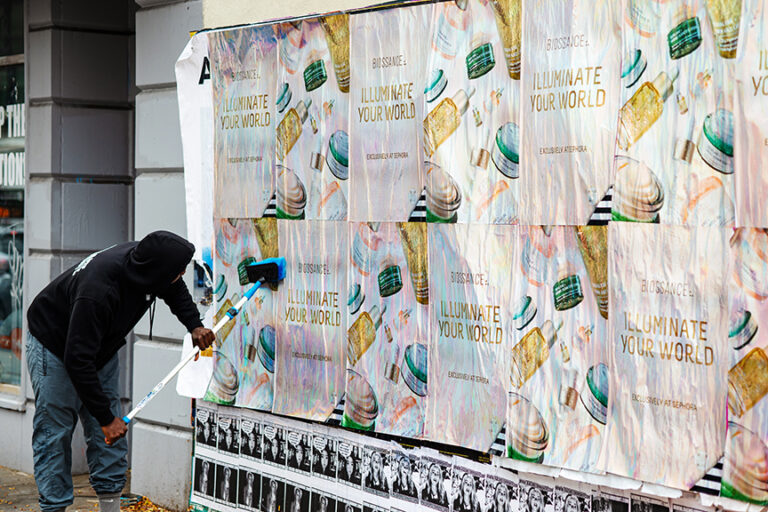 Person applying wheatpaste to a wall covered with custom-printed holographic posters for the Biossance &ldquo;Illuminate Your World&rdquo; campaign, showcasing guerrilla marketing techniques.