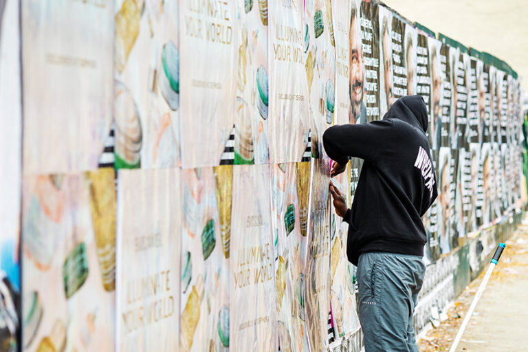 Person applying custom-printed holographic wheat posters for the "Illuminate Your World" campaign by Biossance, showcasing the wheatpaste posting process in an urban setting.