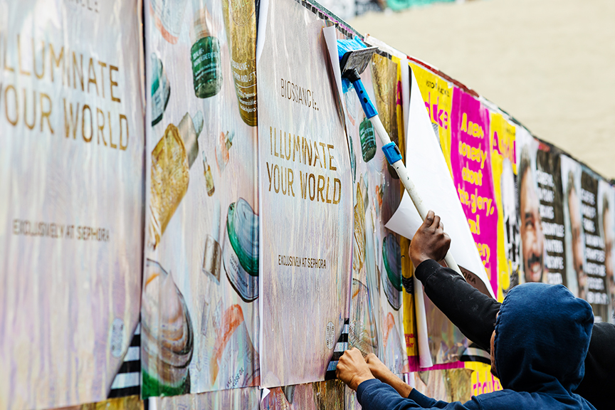 Person applying wheatpaste posters featuring "Illuminate Your World" advertisement in a vibrant urban setting, showcasing guerrilla marketing techniques in Tampa, Florida.