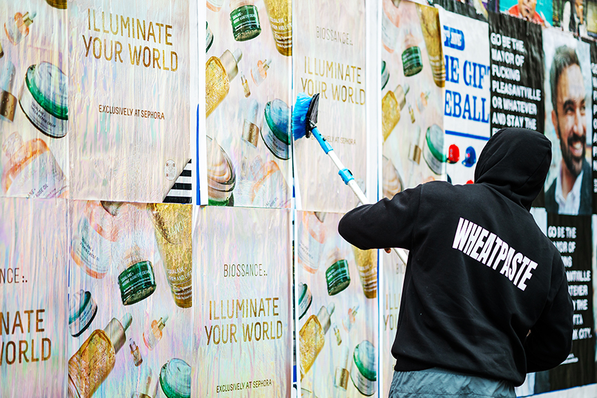 Person applying wheatpaste to a wall covered with colorful posters advertising Biossance's "Illuminate Your World" campaign, showcasing beauty products, in a vibrant urban setting.